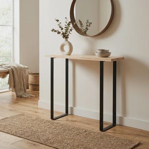 Op de afbeelding: Een rechthoekige sidetable met een licht houten blad en twee zwarte, U-vormige metalen poten. Op de tafel staat een witte, ringvormige keramische vaas met eucalyptus takken en een stapeltje van twee kleine, neutraal gekleurde schaaltjes.