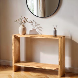 Könnte beinhalten: Eine Konsole aus Naturholz mit einem unteren Regal. Eine beige Vase mit Trockenblumen und ein silberner Schilf-Diffusor stehen auf dem Tisch. Ein schwarz gerahmter Spiegel hängt über dem Tisch. Der Tisch ist etwa 122 cm lang.