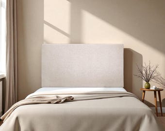 May include: A beige upholstered headboard is the focal point of a bed, complemented by a matching comforter and a folded blanket. The bed is set against a neutral-toned wall, with a small side table and a vase of branches.
