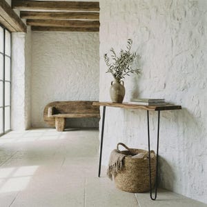 Può includere: Un tavolo console con un piano in legno naturale e sottili gambe a forcina in metallo nero. Sul tavolo si trova un vaso in ceramica con rami verdi e due libri impilati. Un cesto intrecciato con un tessuto sfrangiato si trova sotto.