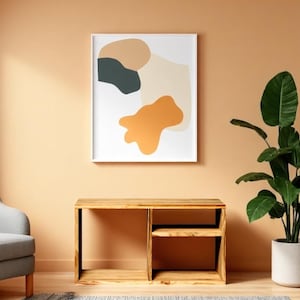 Può includere: Una stampa artistica astratta incorniciata, con forme nei toni del beige, verde scuro e arancione, è appesa sopra una scrivania in legno. A destra c'è una grande pianta in vaso con foglie verdi. A sinistra c'è una poltrona grigia.