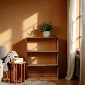 Op de afbeelding: Een houten boekenplank met drie niveaus en een plant in een pot. De boekenplank is gemaakt van lichtkleurig hout. De achtergrond toont een warmgekleurde muur, een ronde houten bijzettafel en een grijze fauteuil. De kamer wordt verlicht door zonlicht.