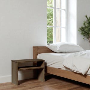 Könnte beinhalten: Ein kleiner Nachttisch aus Holz mit rechteckiger Form und einem Regal. Der Nachttisch ist aus hellbraunem Holz gefertigt und hat ein schlichtes, minimalistisches Design. Er steht neben einem Bett mit weißer Bettwäsche.
