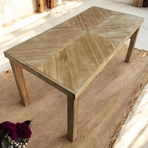 Peut inclure: Table rectangulaire en bois clair avec un motif en chevrons sur le plateau. Elle repose sur quatre pieds robustes et un tapis tissé. Le design général est rustique et naturel, avec des dimensions d'environ 150 cm de long.