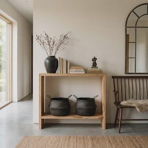 Può includere: Un tavolo consolle in legno chiaro a due ripiani, con un vaso nero contenente rami secchi, libri impilati e una piccola scultura. Due cesti neri intrecciati con manici sono posizionati sul ripiano inferiore, accanto a una panca in legno.