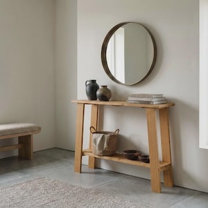 Pode incluir: Uma mesa consola de madeira clara com uma prateleira inferior, com um espelho redondo montado acima. Sobre a mesa estão dois vasos de cerâmica escura e uma pilha de livros. A prateleira inferior contém uma cesta trançada com um pano e duas tigelas escuras.