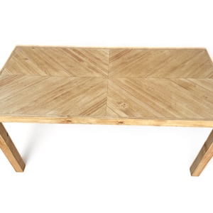 May include: A light brown rectangular wooden table with a geometric pattern on the tabletop. The table has four angled legs and is set against a white background. The tabletop is approximately 60 inches long.
