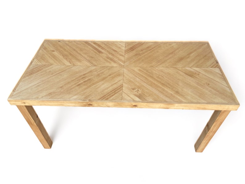 May include: A light brown rectangular wooden table with a geometric pattern on the tabletop. The table has four angled legs and is set against a white background. The tabletop is approximately 60 inches long.