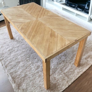 Op de afbeelding: Een rechthoekige houten salontafel met een lichtbruine afwerking. Het tafelblad heeft een visgraatpatroon. De tafel heeft vier stevige poten en staat op een crèmekleurig tapijt. De tafel is ongeveer 122 cm lang en 61 cm breed.