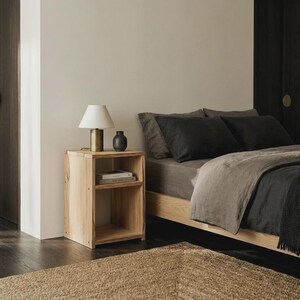 Peut inclure: Une table de chevet en bois clair avec une étagère ouverte, surmontée d'une lampe en laiton avec un abat-jour conique blanc et d'un petit vase gris foncé, placée à côté d'un lit avec une literie gris foncé et un cadre en bois.