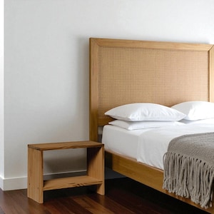 Könnte beinhalten: Ein Nachttisch aus hellem Holz mit einem unteren Regal steht neben einem Bett mit einem gewebten Kopfteil. Das Bett hat weiße Bettwäsche und eine graue Decke mit Fransen. Das Kopfteil ist aus hellem Holz gefertigt.