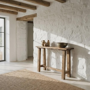 Peut inclure: Une console rectangulaire en bois dotée de quatre pieds et d'une traverse de soutien. Sur le plateau sont posés deux petits vases en céramique beige et un grand bol métallique sombre et peu profond, contre un mur en pierre blanche.