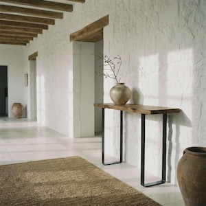 Peut inclure: Une table console rectangulaire en bois avec une finition à grain naturel et deux pieds en métal noir en forme de U. Un vase en céramique ronde de couleur claire contenant une fine branche nue repose sur la surface de la table.