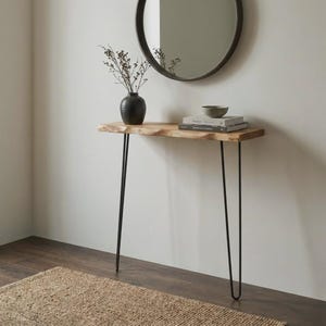 Op de afbeelding: Een sidetable met een natuurlijk houten blad met boomkant en vier dunne zwarte metalen haarspeldpoten. Op de tafel staat een donkere keramische vaas met gedroogde takken en een stapel van twee boeken met daarop een kleine grijze kom.