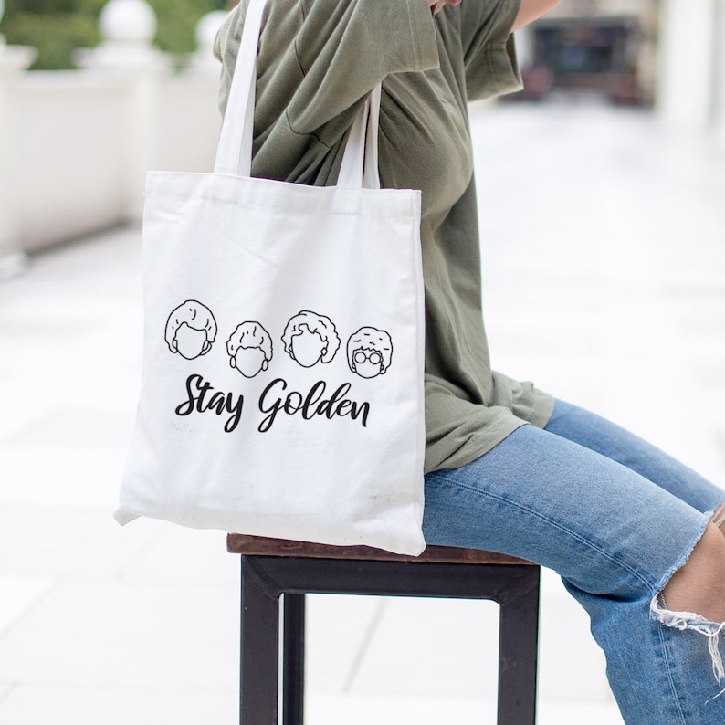 May include: White canvas tote bag with a black outline of four women's faces and the text "Stay Golden".
