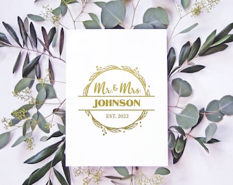 May include: A white card with a gold wreath design and the text "Mr. & Mrs. JOHNSON EST. 2022". The card is surrounded by green eucalyptus branches.