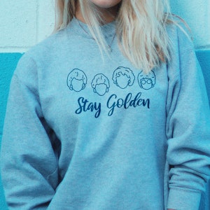 May include: A light blue sweatshirt with a graphic of four women's faces and the text "Stay Golden".