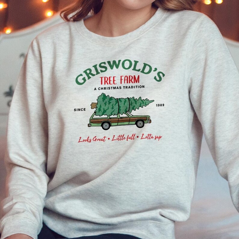 May include: A light gray sweatshirt with the text "GRISWOLD'S TREE FARM" in green, with a station wagon carrying a Christmas tree. The text "A CHRISTMAS TRADITION" and "SINCE 1989" are also printed on the sweatshirt. The phrase "Looks Great &bull; Little full &bull; Lotta sap" is printed below the car.
