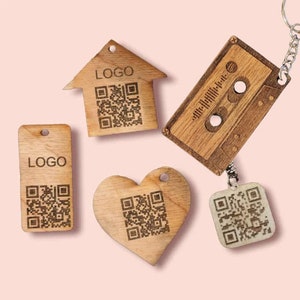 May include: Five wooden keychains with different shapes and designs. Each keychain features a unique QR code and the word "LOGO" engraved on the wood. The keychains are in the shapes of a house, a heart, a cassette tape, and two rectangular tags.