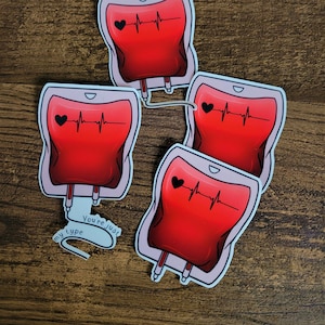 Op de afbeelding: Vier rode bloedzak-stickers met een zwart hart en een hartslaglijn. De stickers hebben een witte omtrek en bevinden zich op een houten oppervlak. Eén sticker heeft de tekst "You're just my type" op een wit lint.