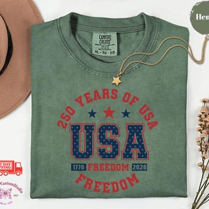 May include: A sage green t-shirt with the text "250 YEARS OF USA" in red and blue. The shirt features the words "FREEDOM" and "1776 FREEDOM 2026". A gold necklace with a star pendant is on the shirt.
