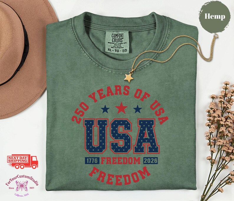May include: A sage green t-shirt with the text "250 YEARS OF USA" in red and blue. The shirt features the words "FREEDOM" and "1776 FREEDOM 2026". A gold necklace with a star pendant is on the shirt.