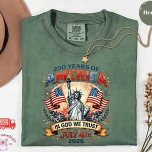 May include: A green t-shirt with a graphic celebrating 250 years of America. The design includes the Statue of Liberty, American flags, and the text "In God We Trust" and "July 4th 2026". A gold necklace with a star pendant is draped across the shirt.
