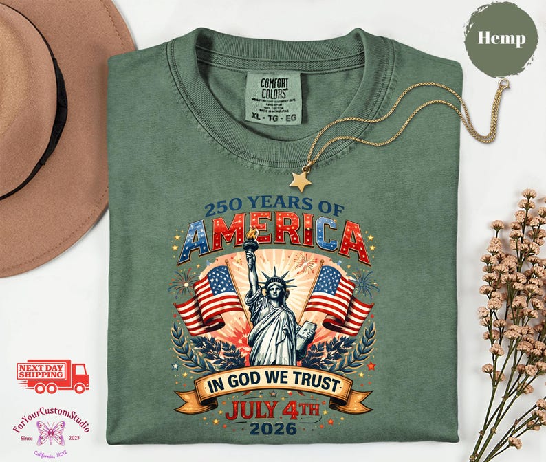 May include: A green t-shirt with a graphic celebrating 250 years of America. The design includes the Statue of Liberty, American flags, and the text "In God We Trust" and "July 4th 2026". A gold necklace with a star pendant is draped across the shirt.