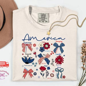 May include: Cream-colored t-shirt with the word "America" in blue script. The shirt features patriotic bow designs, flowers, and inspirational words. A gold star pendant necklace and a brown hat are also visible.