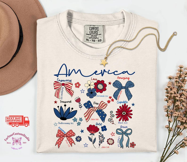 May include: Cream-colored t-shirt with the word "America" in blue script. The shirt features patriotic bow designs, flowers, and inspirational words. A gold star pendant necklace and a brown hat are also visible.