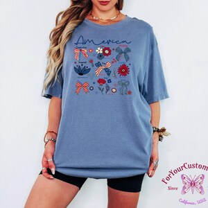 May include: Blue t-shirt with the word "America" in script. The shirt features patriotic bow and floral graphics in red, white, and blue. The shirt is a comfortable, oversized fit.