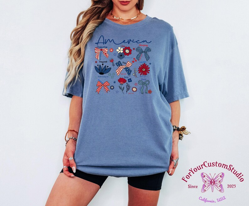May include: Blue t-shirt with the word "America" in script. The shirt features patriotic bow and floral graphics in red, white, and blue. The shirt is a comfortable, oversized fit.