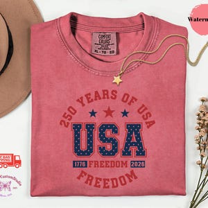 May include: A watermelon-colored t-shirt with the text "250 YEARS OF USA" in red and blue. The word "USA" is prominently displayed. A gold star necklace rests on the shirt. The shirt also features the text "1776 FREEDOM 2026 FREEDOM".