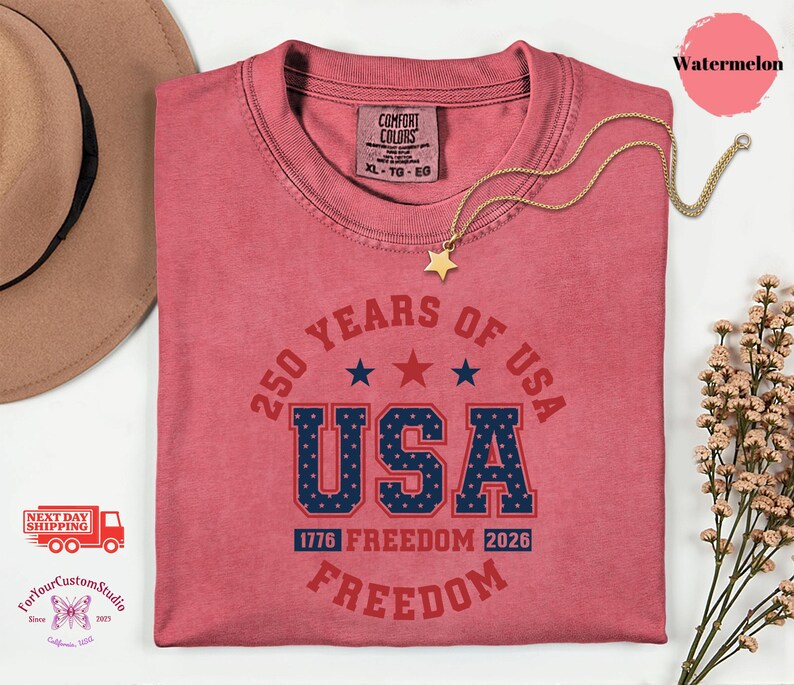 May include: A watermelon-colored t-shirt with the text "250 YEARS OF USA" in red and blue. The word "USA" is prominently displayed. A gold star necklace rests on the shirt. The shirt also features the text "1776 FREEDOM 2026 FREEDOM".