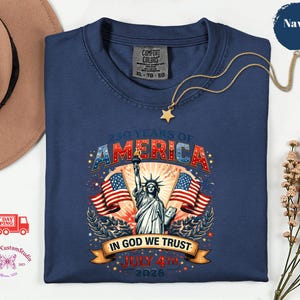 May include: Navy blue t-shirt with a graphic celebrating 250 years of America. The design features the Statue of Liberty, American flags, and the text "IN GOD WE TRUST" and "JULY 4TH 2026". A gold star necklace is draped across the shirt.