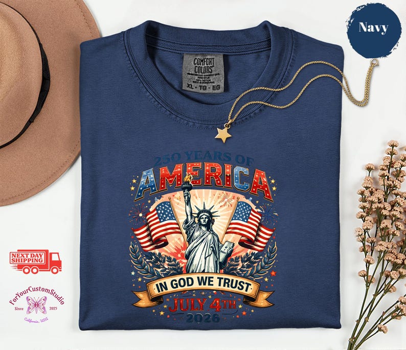 May include: Navy blue t-shirt with a graphic celebrating 250 years of America. The design features the Statue of Liberty, American flags, and the text "IN GOD WE TRUST" and "JULY 4TH 2026". A gold star necklace is draped across the shirt.