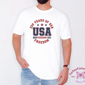 May include: White t-shirt featuring a patriotic design celebrating 250 years of the USA. The design includes the word "USA" in blue with a star pattern, red stars, and the words "Freedom" and "1776 Freedom 2026".