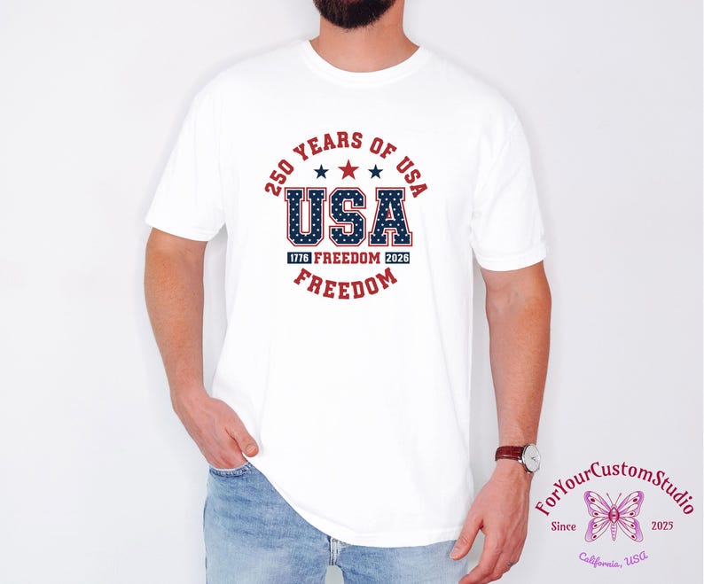 May include: White t-shirt featuring a patriotic design celebrating 250 years of the USA. The design includes the word "USA" in blue with a star pattern, red stars, and the words "Freedom" and "1776 Freedom 2026".