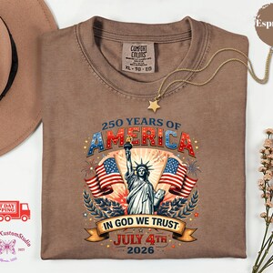 May include: Espresso-colored t-shirt with a graphic celebrating 250 years of America. The design includes the Statue of Liberty, American flags, and the text "In God We Trust" and "July 4th 2026". The shirt has a Comfort Colors tag.