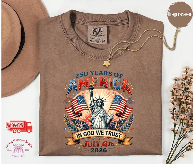 May include: Espresso-colored t-shirt with a graphic celebrating 250 years of America. The design includes the Statue of Liberty, American flags, and the text "In God We Trust" and "July 4th 2026". The shirt has a Comfort Colors tag.