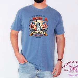 May include: A blue t-shirt featuring a graphic celebrating 250 years of America. The design includes the Statue of Liberty, American flags, and the text "IN GOD WE TRUST" and "JULY 4TH 2026". The shirt is a comfortable fit.