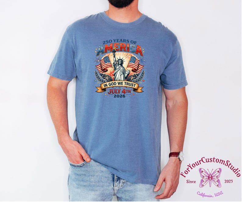 May include: A blue t-shirt featuring a graphic celebrating 250 years of America. The design includes the Statue of Liberty, American flags, and the text "IN GOD WE TRUST" and "JULY 4TH 2026". The shirt is a comfortable fit.