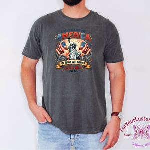 May include: Dark gray t-shirt featuring a patriotic design celebrating 250 years of America. The graphic includes the Statue of Liberty, American flags, and the text "In God We Trust" and "July 4th 2026".