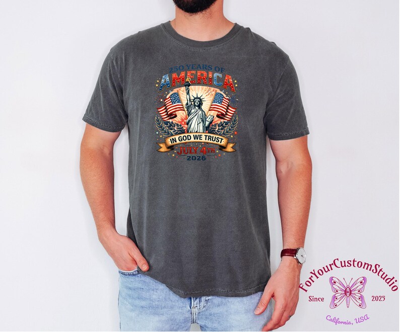 May include: Dark gray t-shirt featuring a patriotic design celebrating 250 years of America. The graphic includes the Statue of Liberty, American flags, and the text "In God We Trust" and "July 4th 2026".