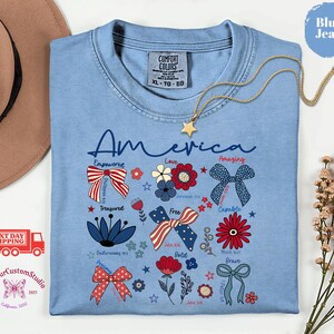 May include: A light blue t-shirt with the word "America" in script, decorated with patriotic bows, flowers, and text. A gold star necklace hangs on the shirt. The shirt has the Comfort Colors brand tag.