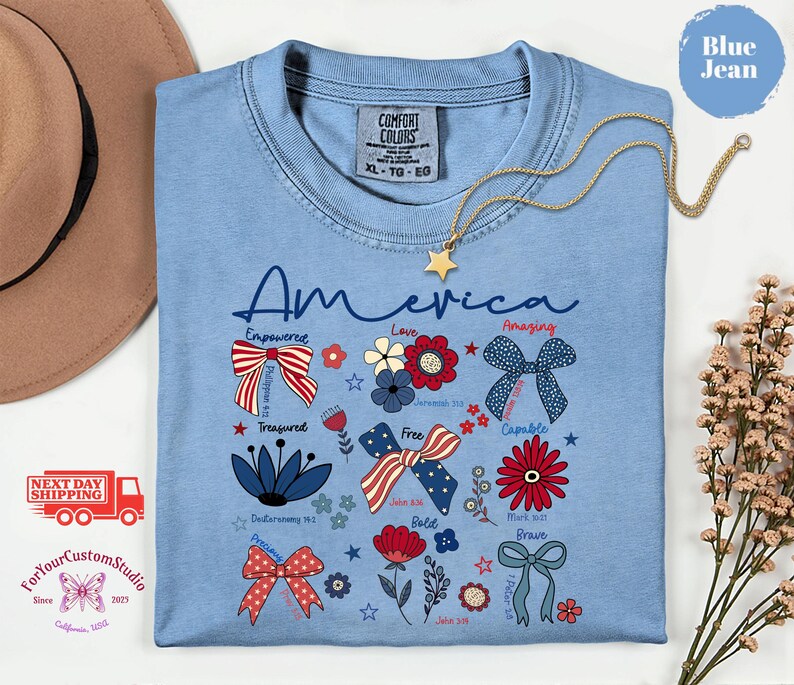 May include: A light blue t-shirt with the word "America" in script, decorated with patriotic bows, flowers, and text. A gold star necklace hangs on the shirt. The shirt has the Comfort Colors brand tag.