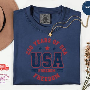 May include: Navy blue t-shirt with the text "250 YEARS OF USA" in red, with the word "USA" in a bold, patriotic font. The shirt also features the text "1776 FREEDOM 2026 FREEDOM". A gold necklace with a star pendant is on the shirt.