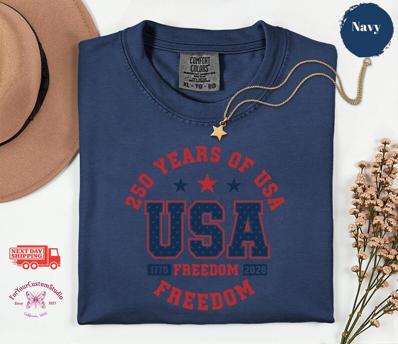 May include: Navy blue t-shirt with the text "250 YEARS OF USA" in red, with the word "USA" in a bold, patriotic font. The shirt also features the text "1776 FREEDOM 2026 FREEDOM". A gold necklace with a star pendant is on the shirt.