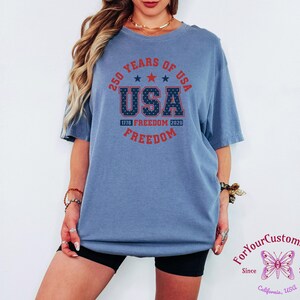 May include: A blue short-sleeved t-shirt with the text "250 YEARS OF USA" in a circular design. The word "USA" is prominently displayed with a star pattern, and "FREEDOM" is written below. The shirt commemorates 1776-2026.