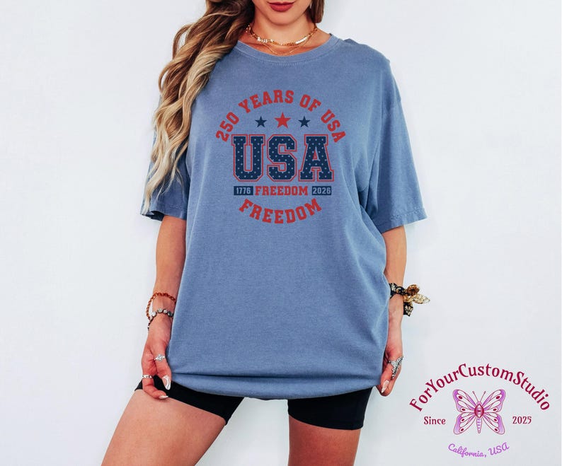 May include: A blue short-sleeved t-shirt with the text "250 YEARS OF USA" in a circular design. The word "USA" is prominently displayed with a star pattern, and "FREEDOM" is written below. The shirt commemorates 1776-2026.